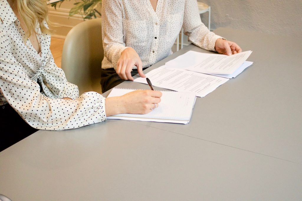woman signing on white printer paper beside woman about to touch the documents paperlass